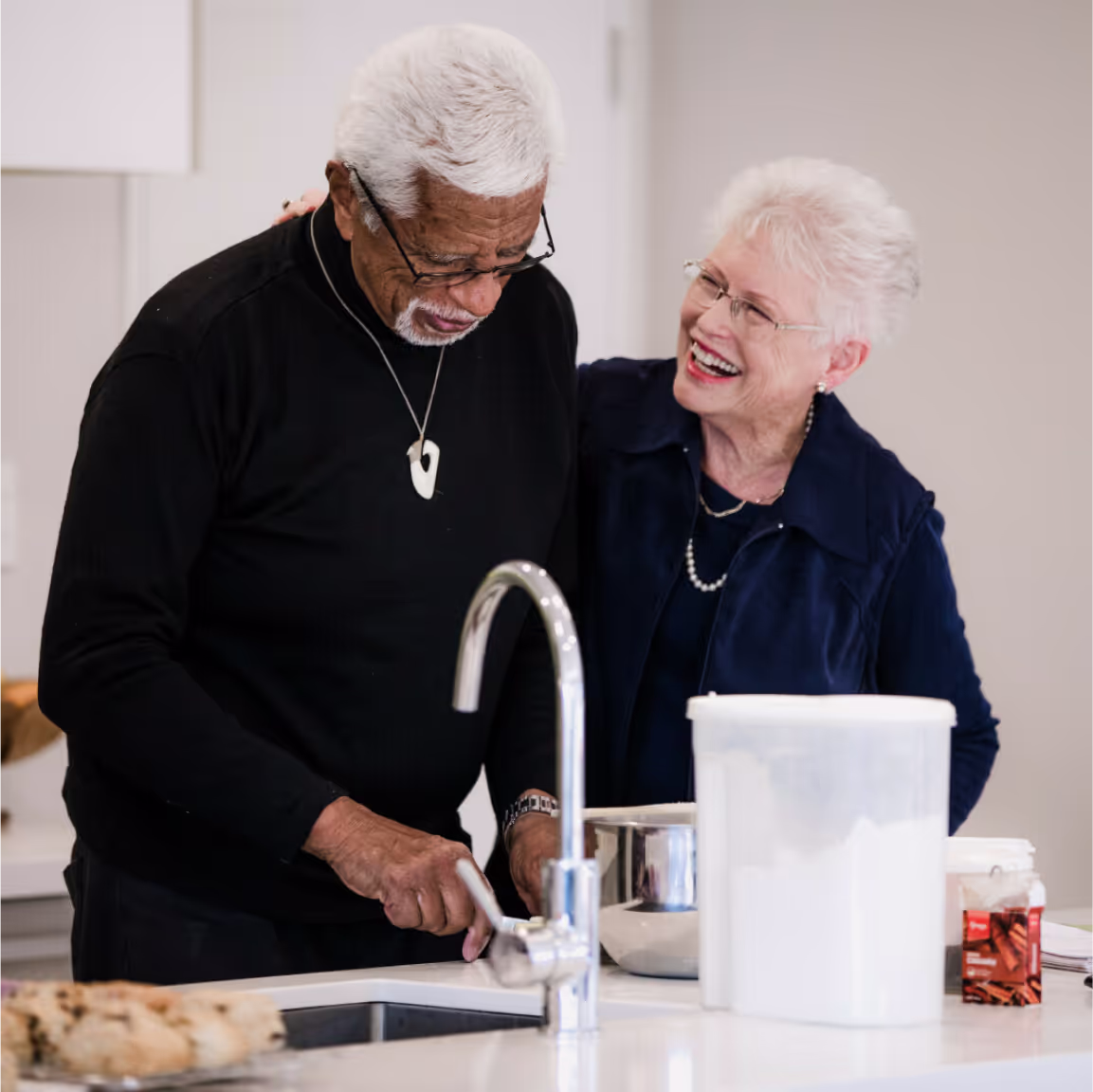 An older couple, both wearing glasses, interact while they're standing in a kitchen